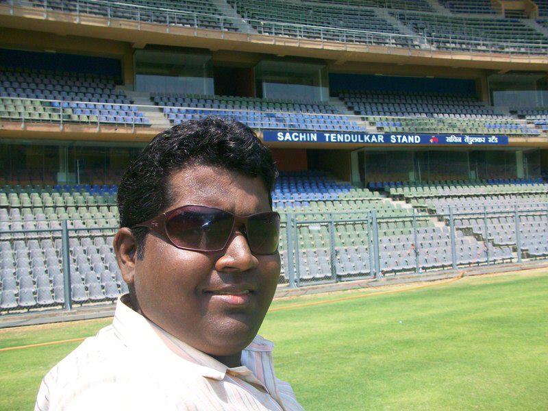 A man poses at Wankhede Stadium with the Sachin Tendulkar Stand visible, reflecting India's passion for cricket.