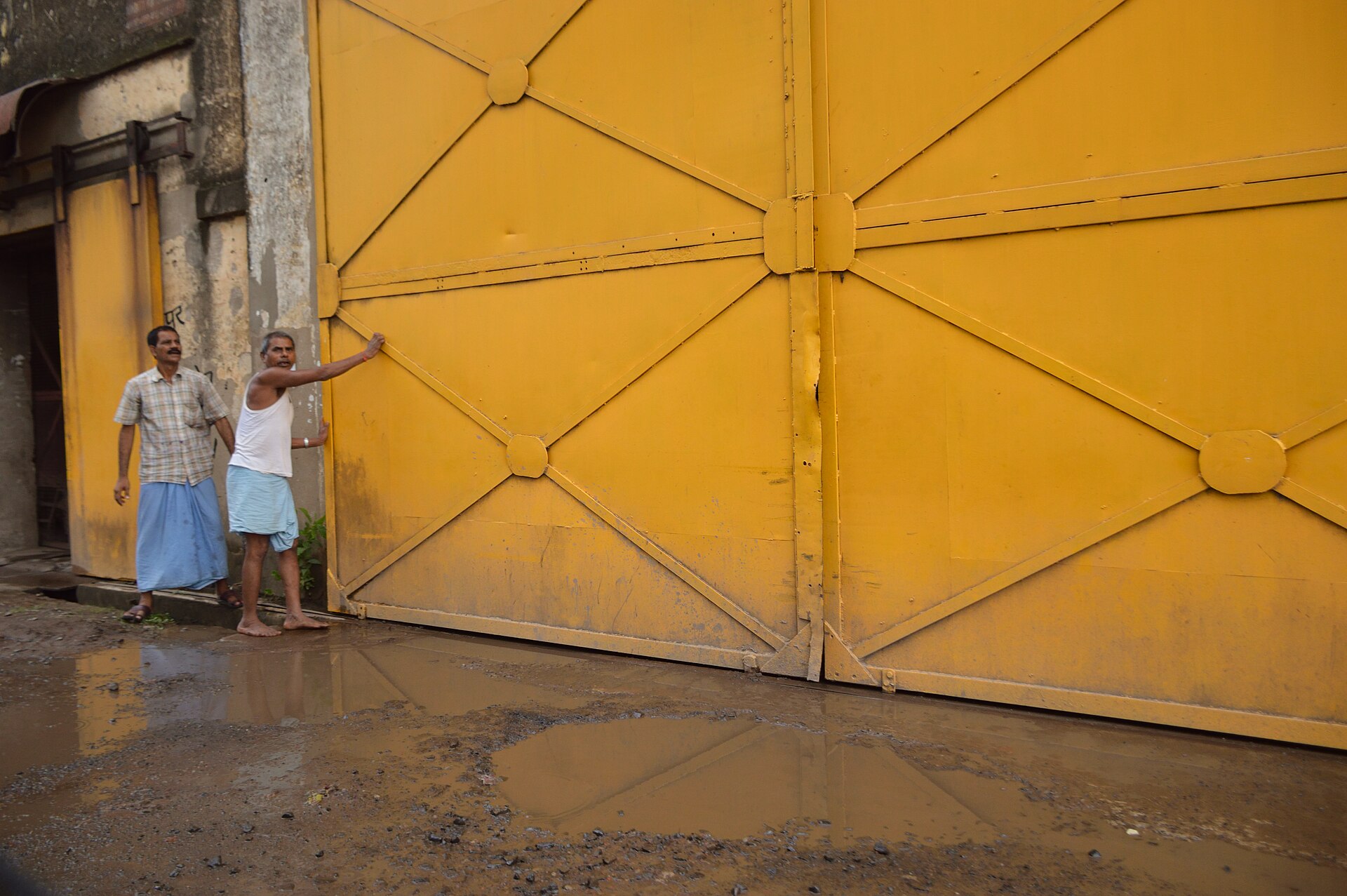 Jute Mill Gate | Indian Press Two men stand near a large yellow gate at an India jute mill, showing the impact of flooding on the economy.