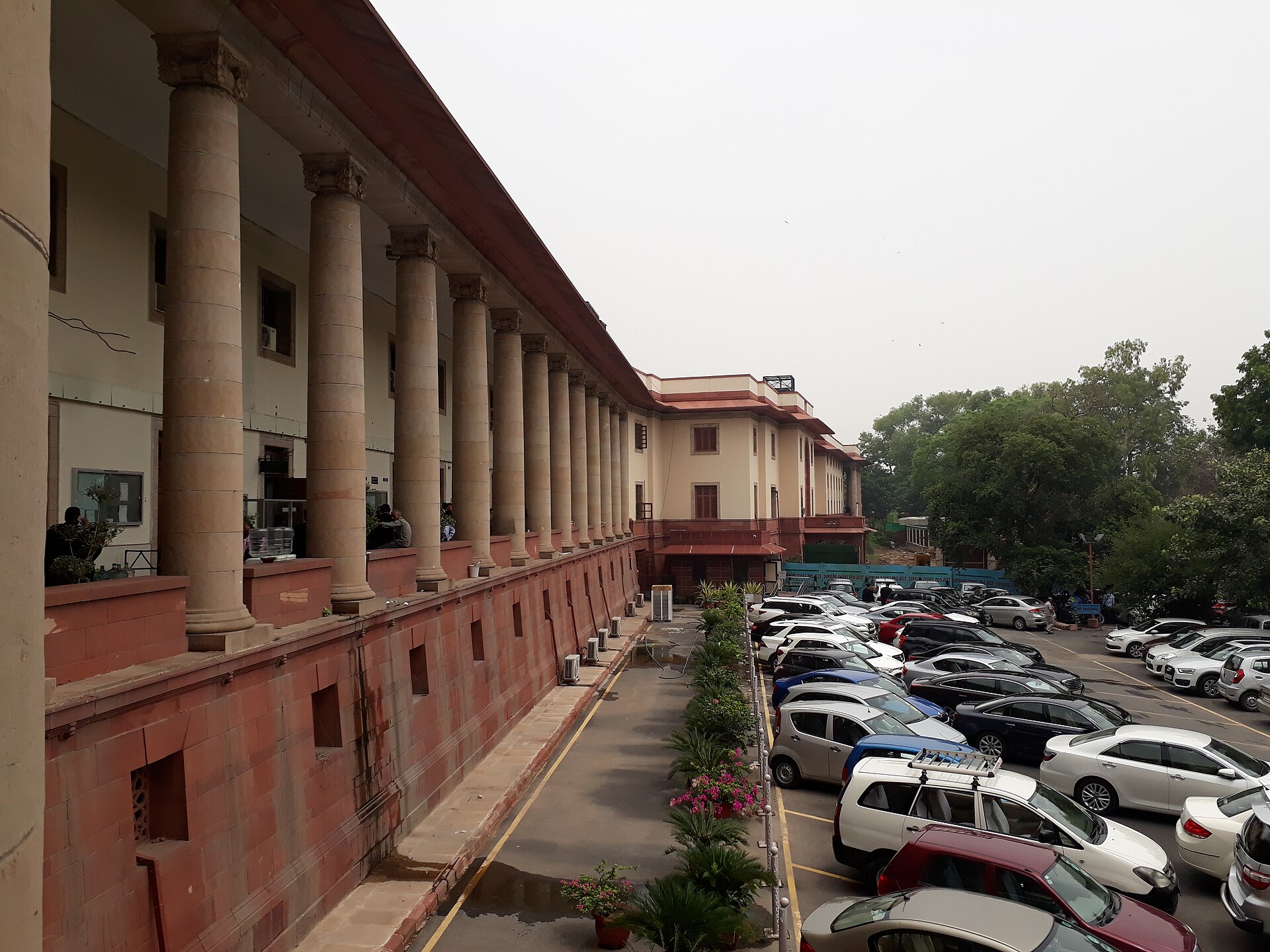 Supreme Court of India | Indian Press A long government building with columns overlooks a parking lot filled with cars in New Delhi.